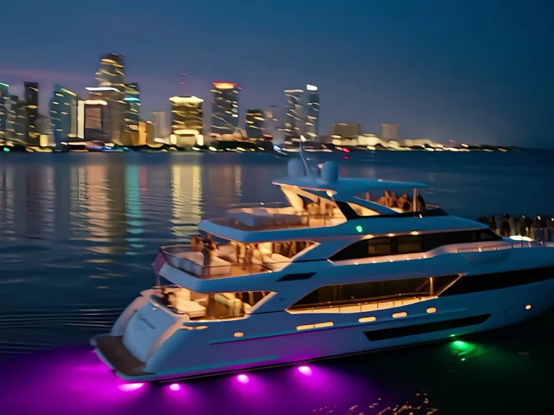 A white yacht lit in purple on the water against a night city skyline