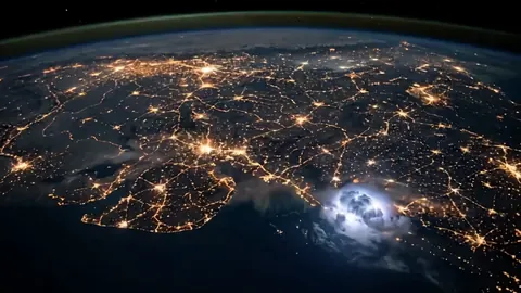 Night view of the Indian subcontinent from orbit, cities glowing gold, a lightning flash at the lower right.