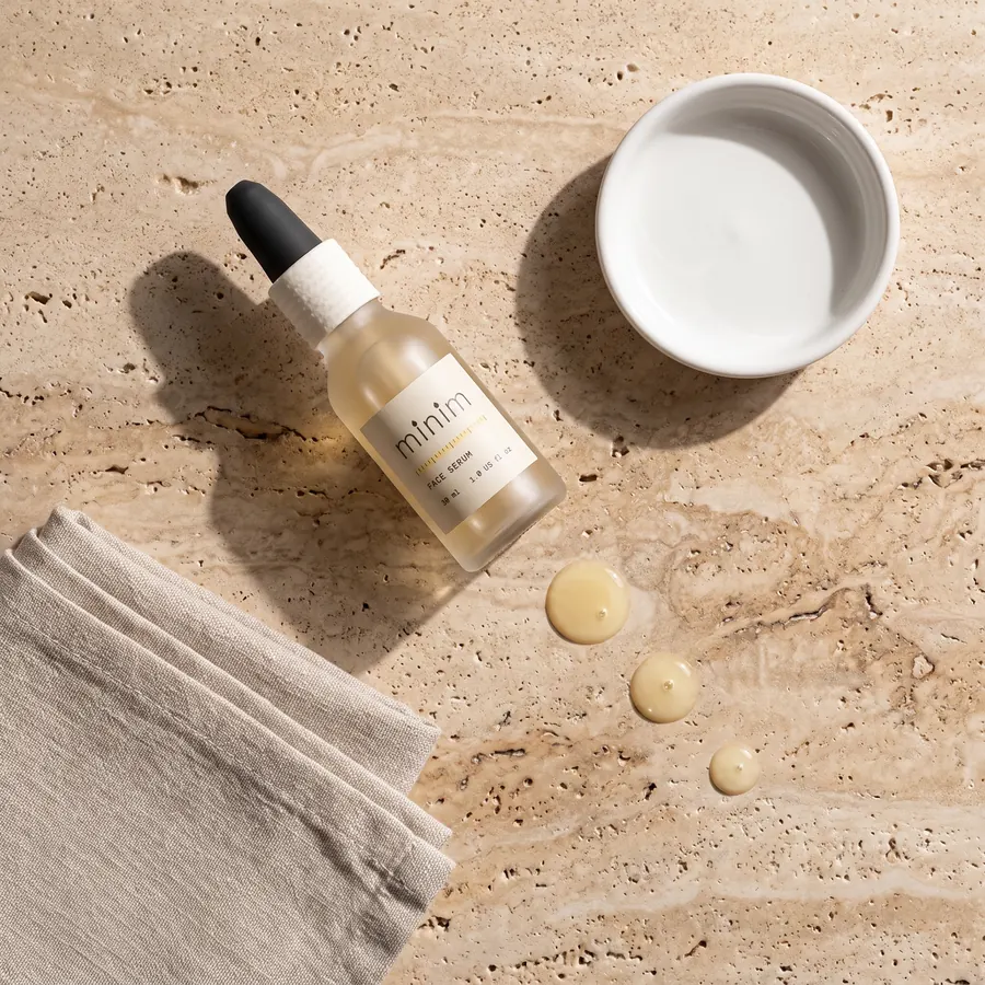 Flat lay on pale travertine: the serum bottle, a white dish, a folded linen cloth and exactly three golden serum drops.