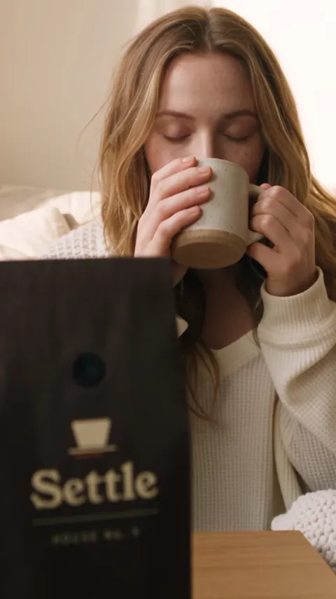 A woman sipping from a stoneware mug, the coffee bag blurred in the foreground.