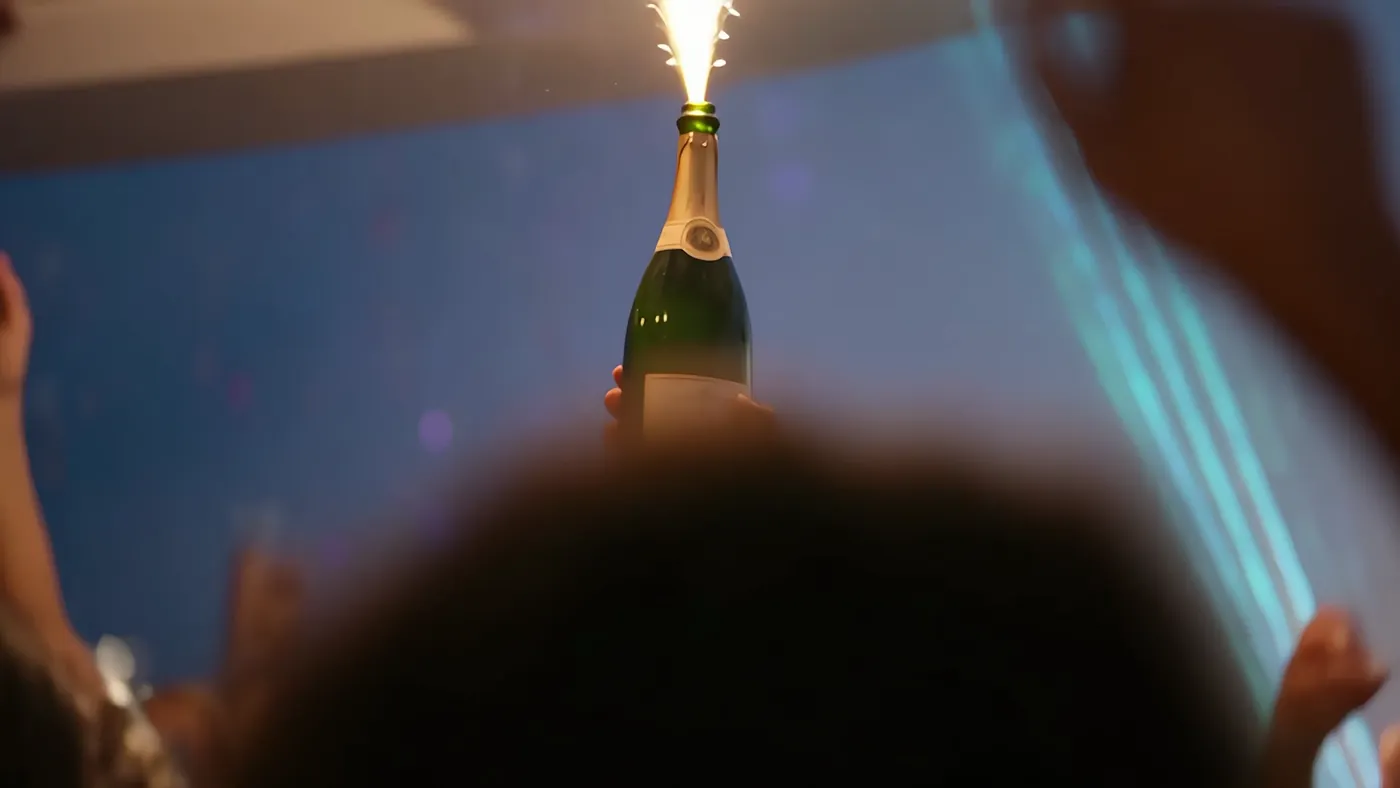 A champagne bottle held up with a lit sparkler, teal light streaking behind and silhouettes below.