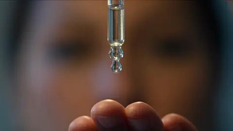 Macro shot of a glass dropper with one serum drop about to fall, her blurred face behind it.