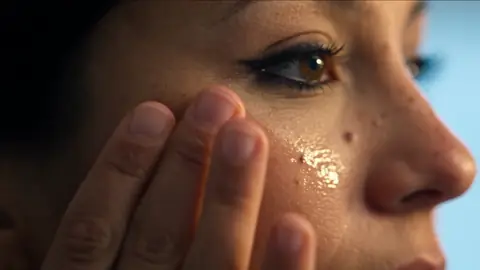 Close portrait of her eye and cheek as she presses a fingertip to her skin, a glossy serum drop on her cheekbone.