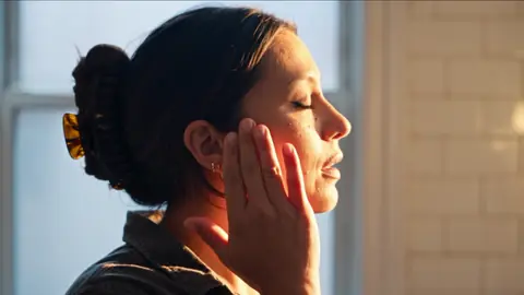 Profile of her face in warm window light, eyes closed, hand resting against her cheek in relief.