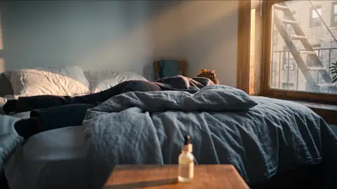 A bedroom in blue evening light: she lies asleep under a linen duvet, the small serum bottle on the bedside table.