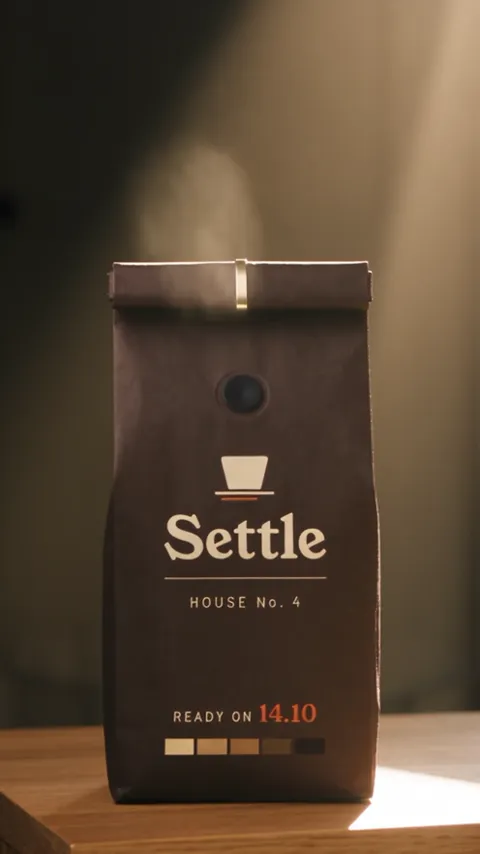 A dark roasted-coffee bag standing on a wooden shelf in low sun, a soft puff of aroma lifting from its folded top.