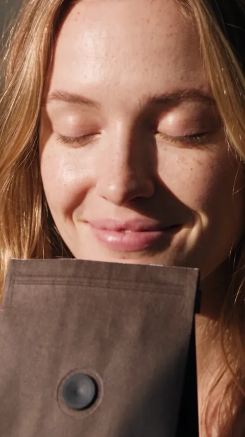 Close portrait of a woman with freckles and her eyes closed, smiling as she breathes in, a coffee bag at the bottom of the frame.