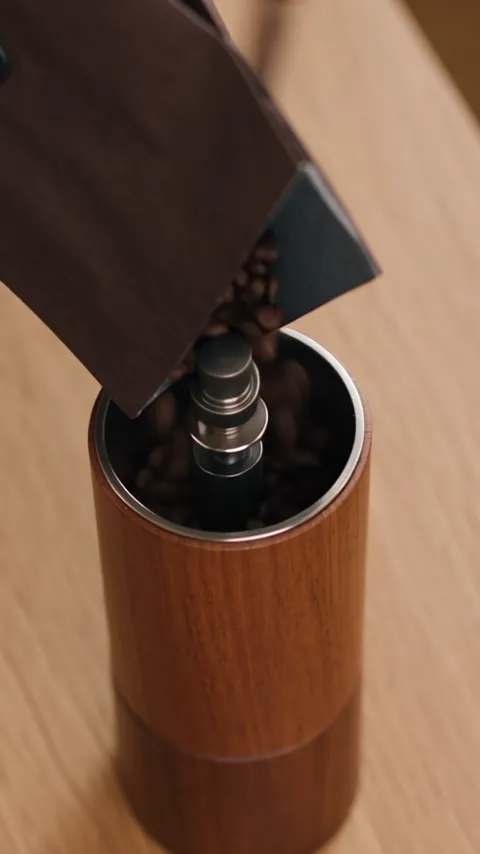 Roasted beans tipping from a bag into a wooden hand grinder.