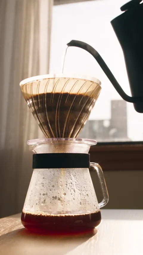 A kettle pouring into a glass pour-over dripper over a carafe, window light behind.